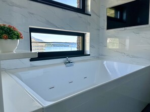 A modern bathroom with a large white bathtub, a window with a view of a building and water, and a potted plant on the counter.