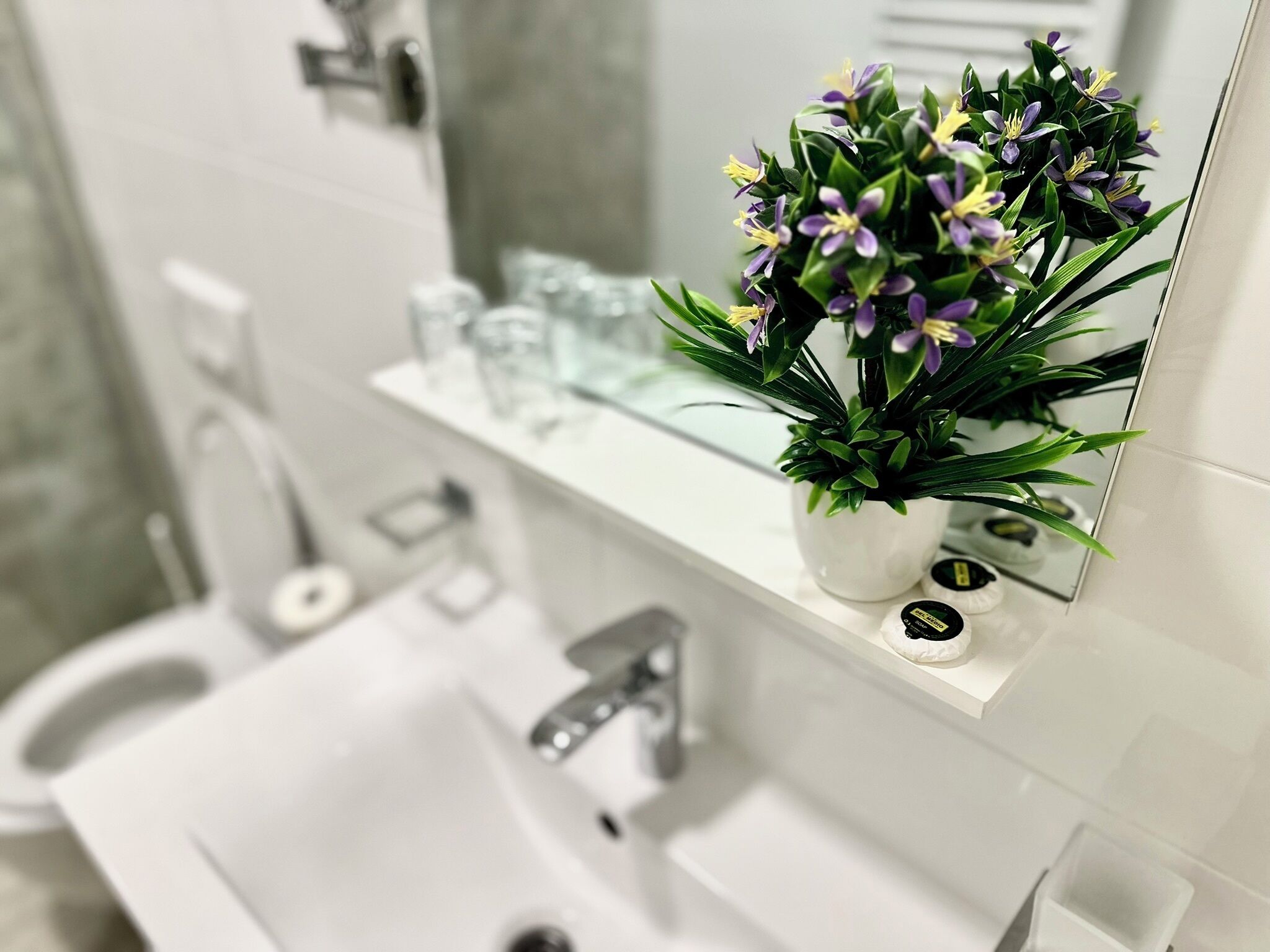 Une salle de bain avec un lavabo blanc, un miroir et une plante en pot aux fleurs violettes.