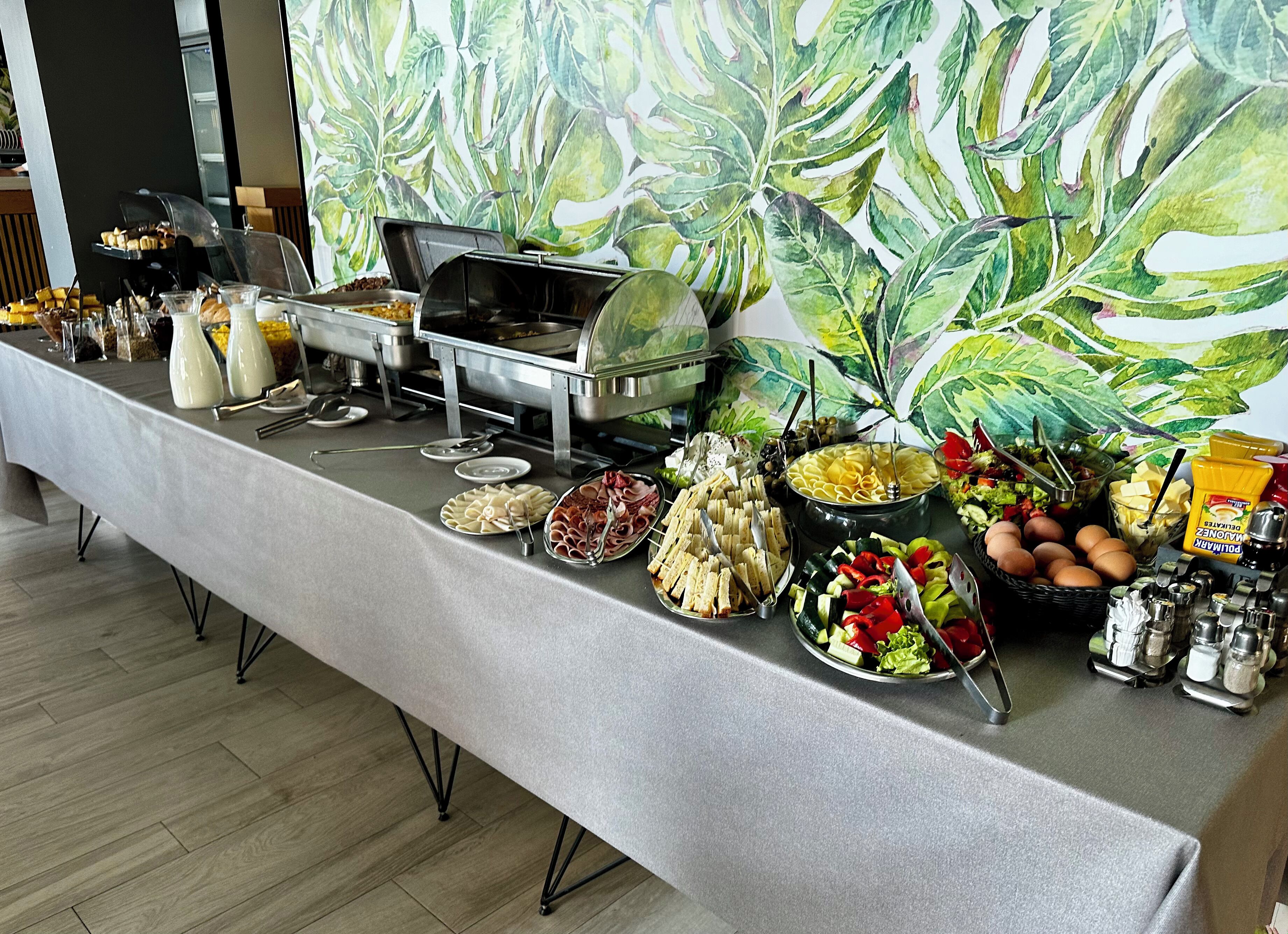 En buffet med forskellige retter og tilbehør foran en stor murals med frodige planter.