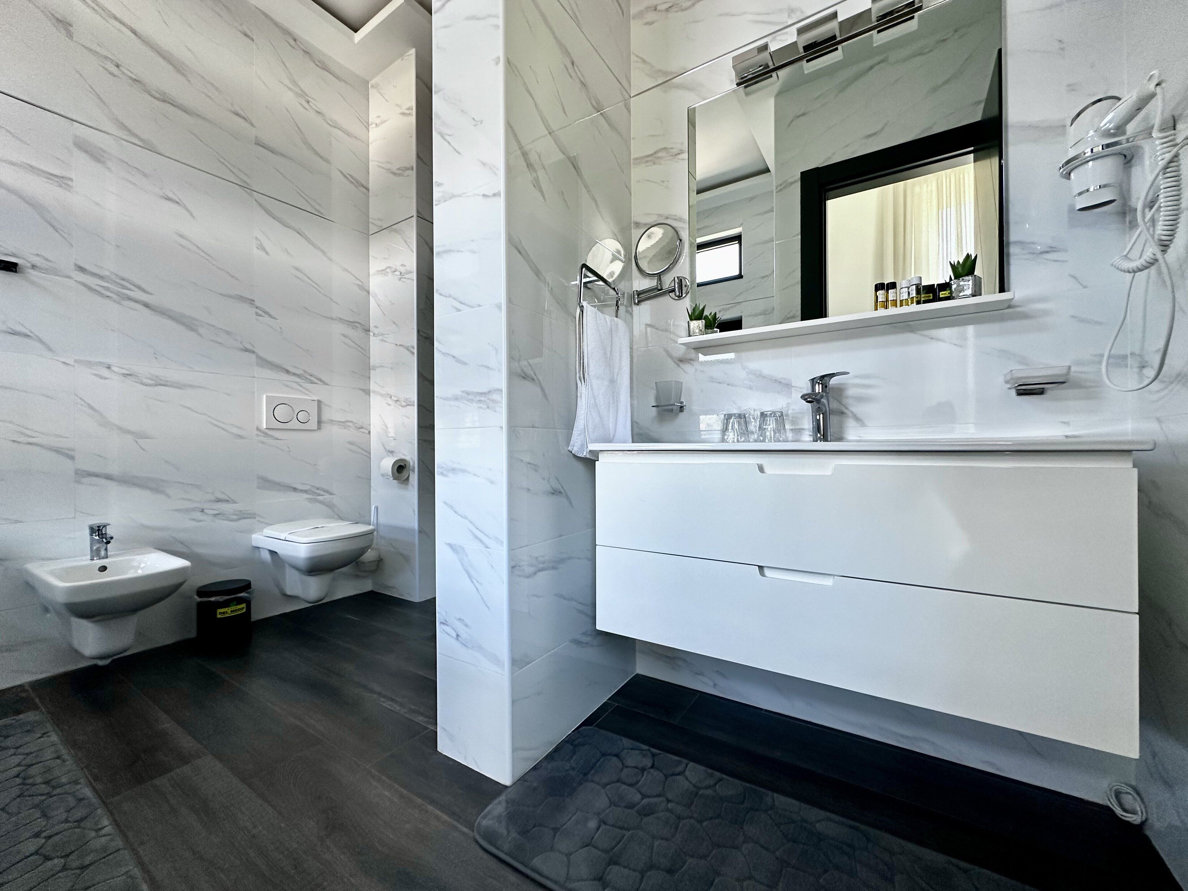 A modern bathroom with a marble wall, a white vanity, a bidet, and a shower area.