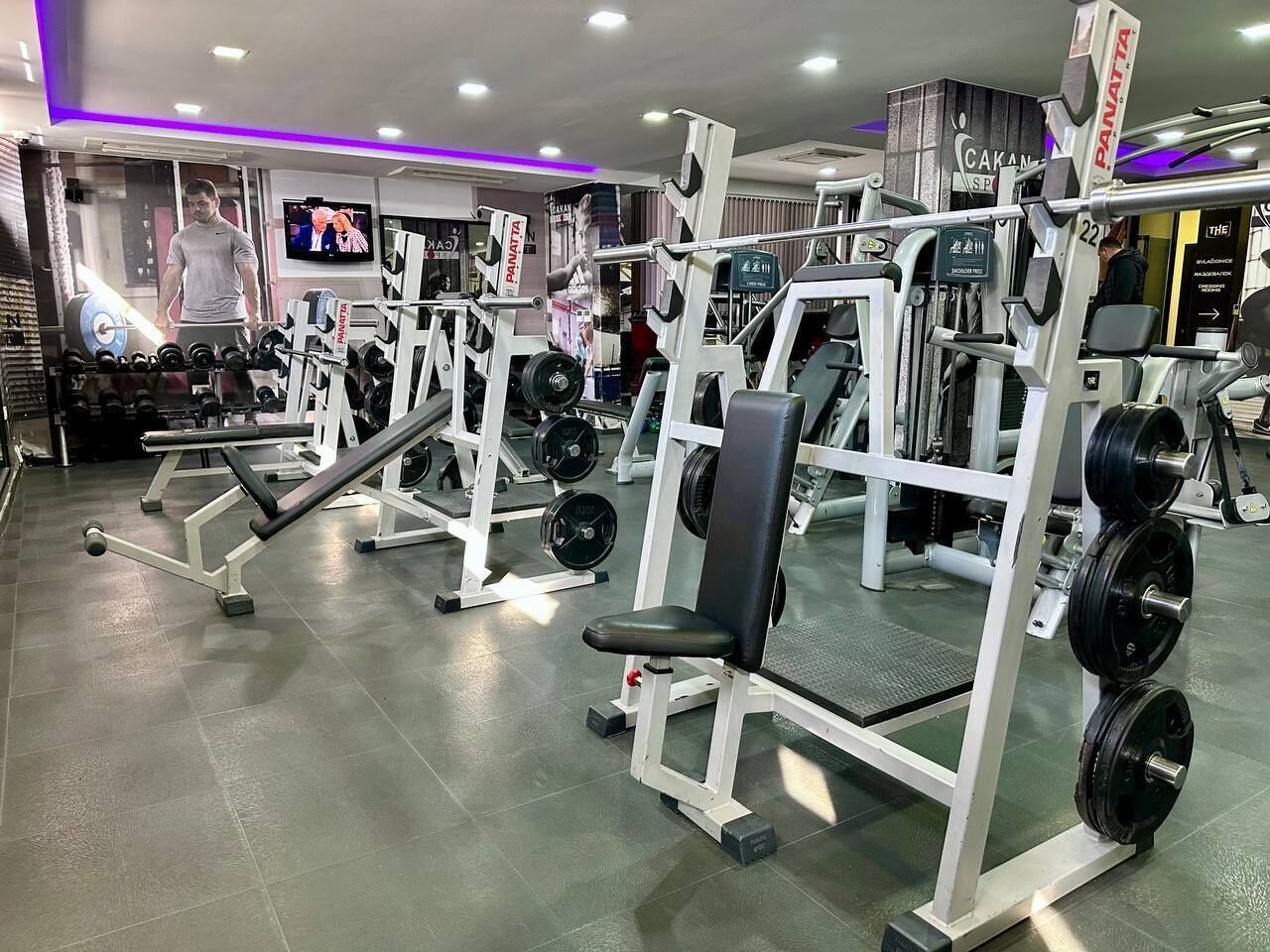 Un gimnasio con equipamiento de levantamiento de pesas variado, incluyendo bancos y estructuras para sentadillas, y un televisor montado en la pared.
