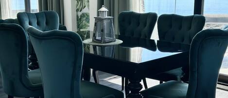 A dining area with a glass table surrounded by teal chairs, a lantern, and a view of the sea.