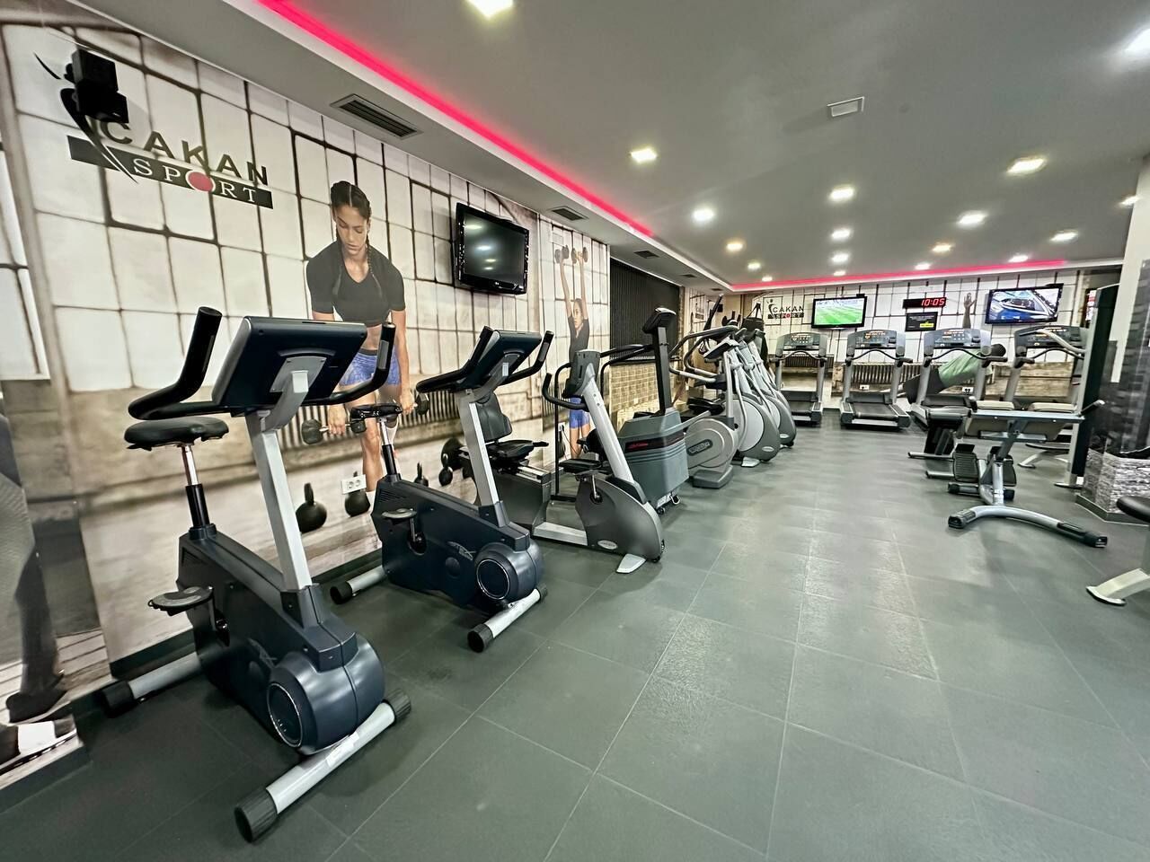 Un gimnasio con diversas máquinas de cardio, un televisor montado en la pared y un reloj digital que marca las 10:05.