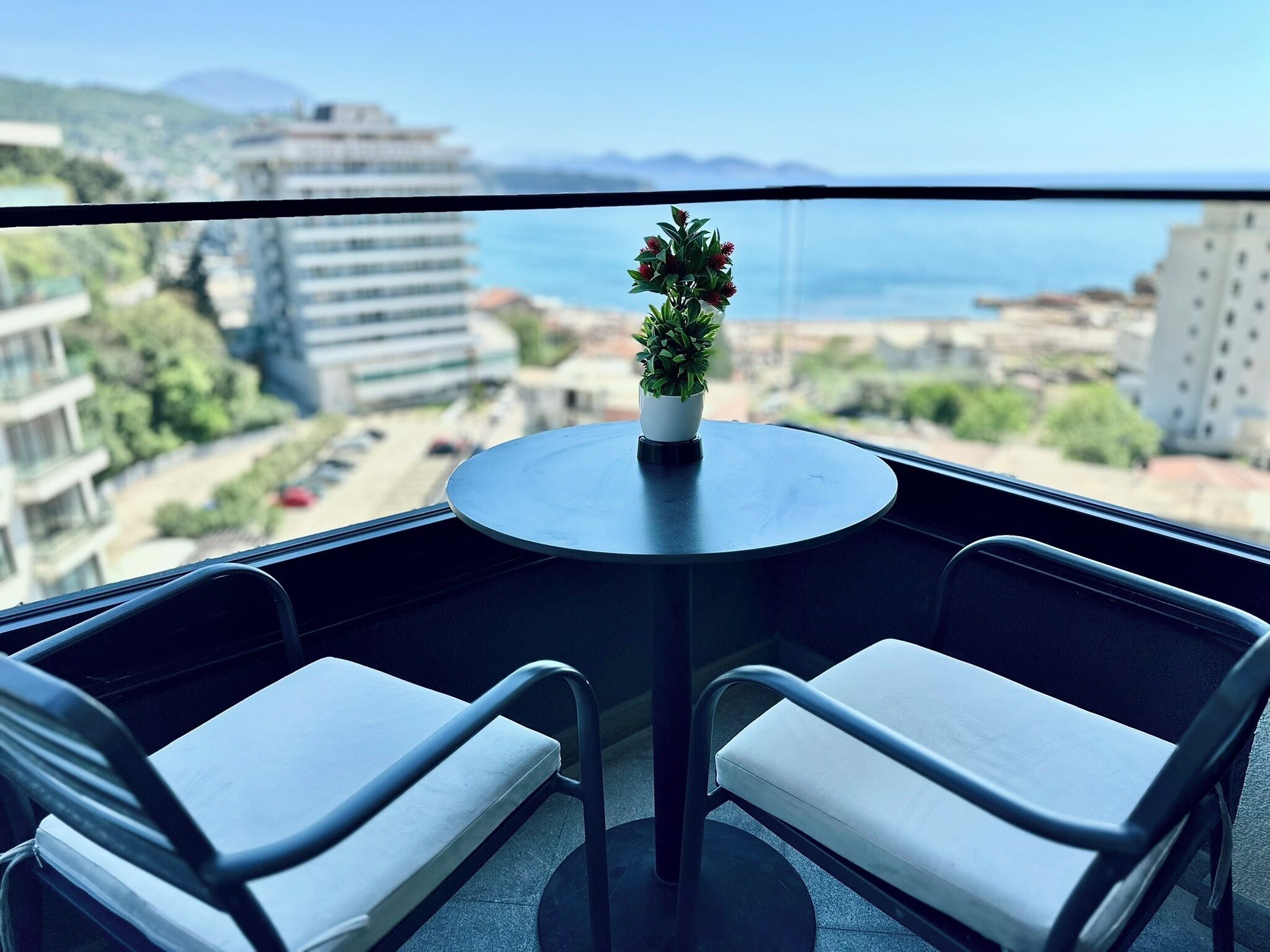 A balcony with a round table and two chairs, overlooking a cityscape with buildings and a body of water.