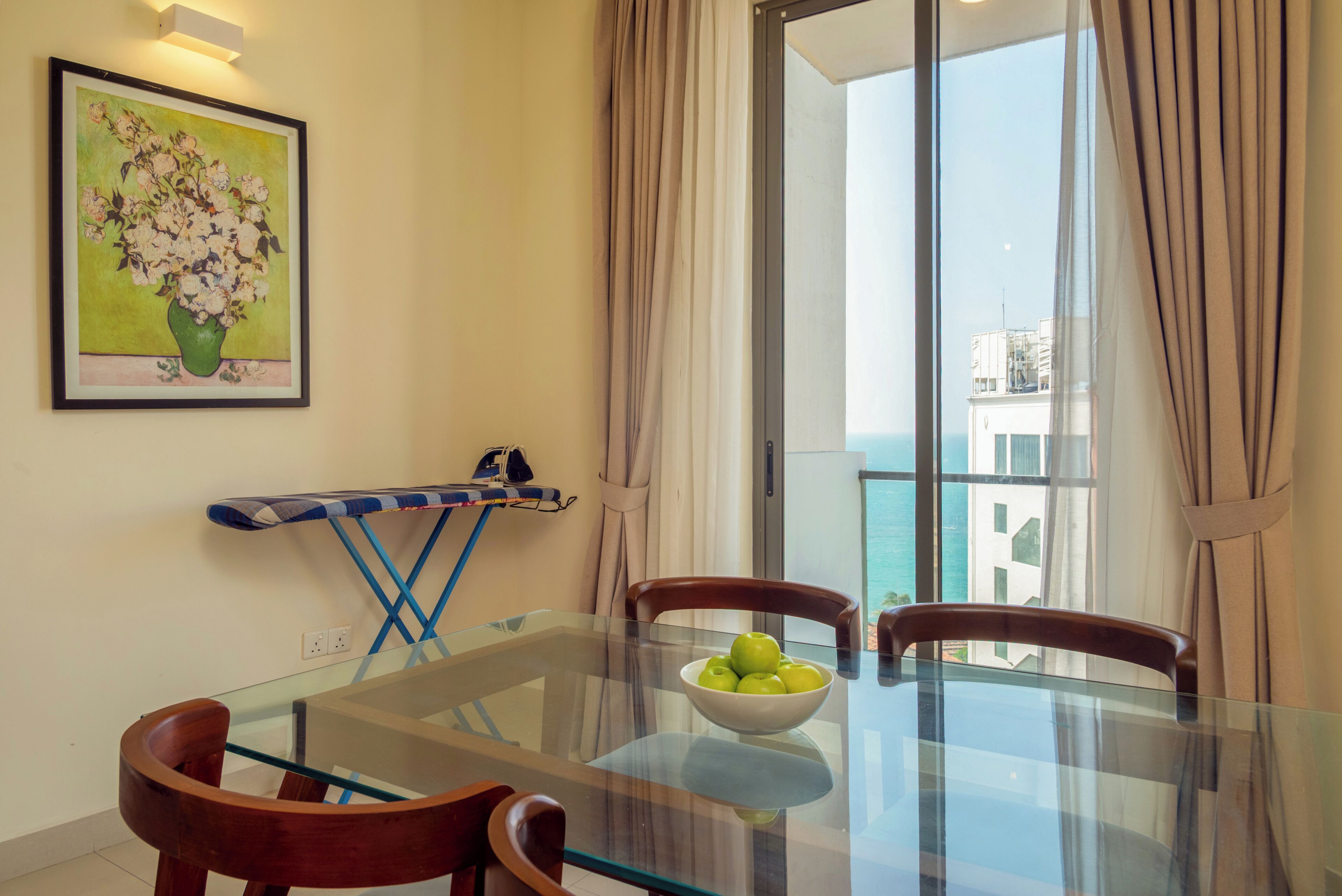 A dining area with a glass table, wooden chairs, a bowl of apples, a painting on the wall, and a view of the sea through a large window.