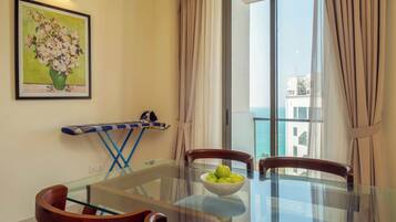 A dining area with a glass table, wooden chairs, a bowl of apples, a painting on the wall, and a view of the sea through a large window.