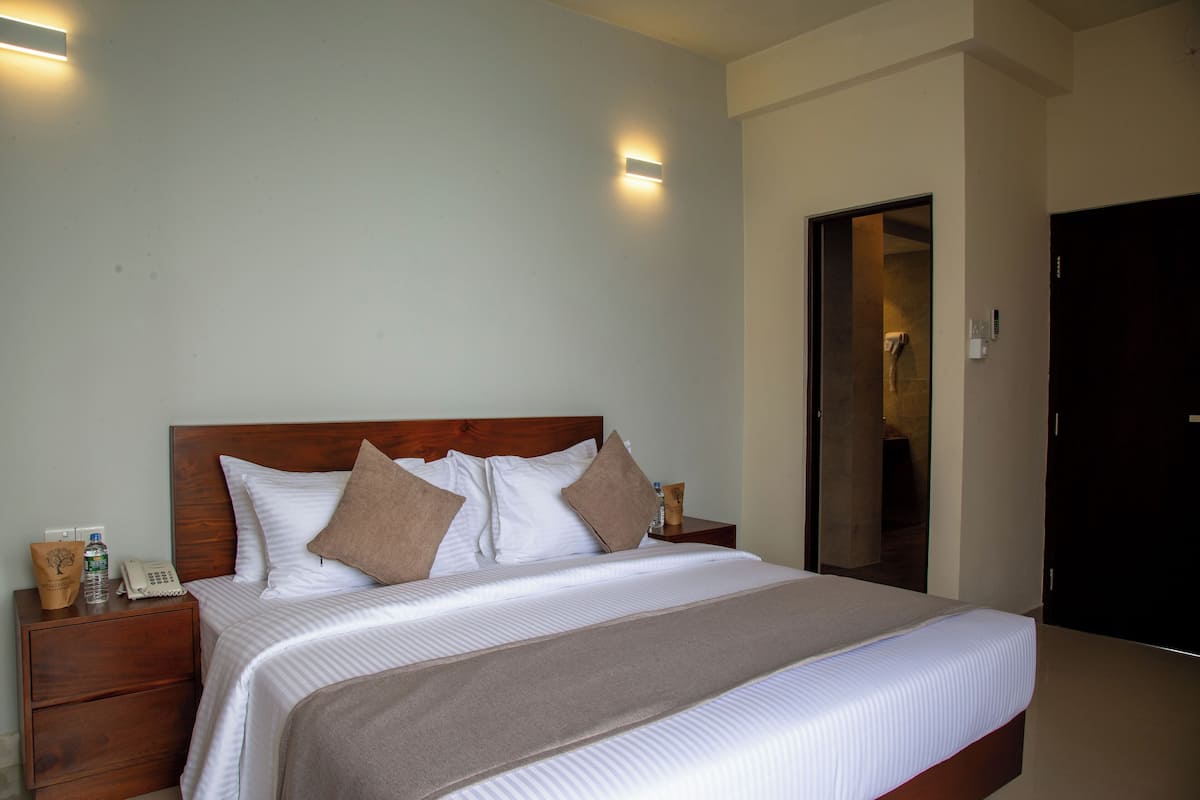 Cama bem arrumada com cabeceira de madeira, mesa de cabeceira com telefone e um frasco, e luminária fixada na parede.