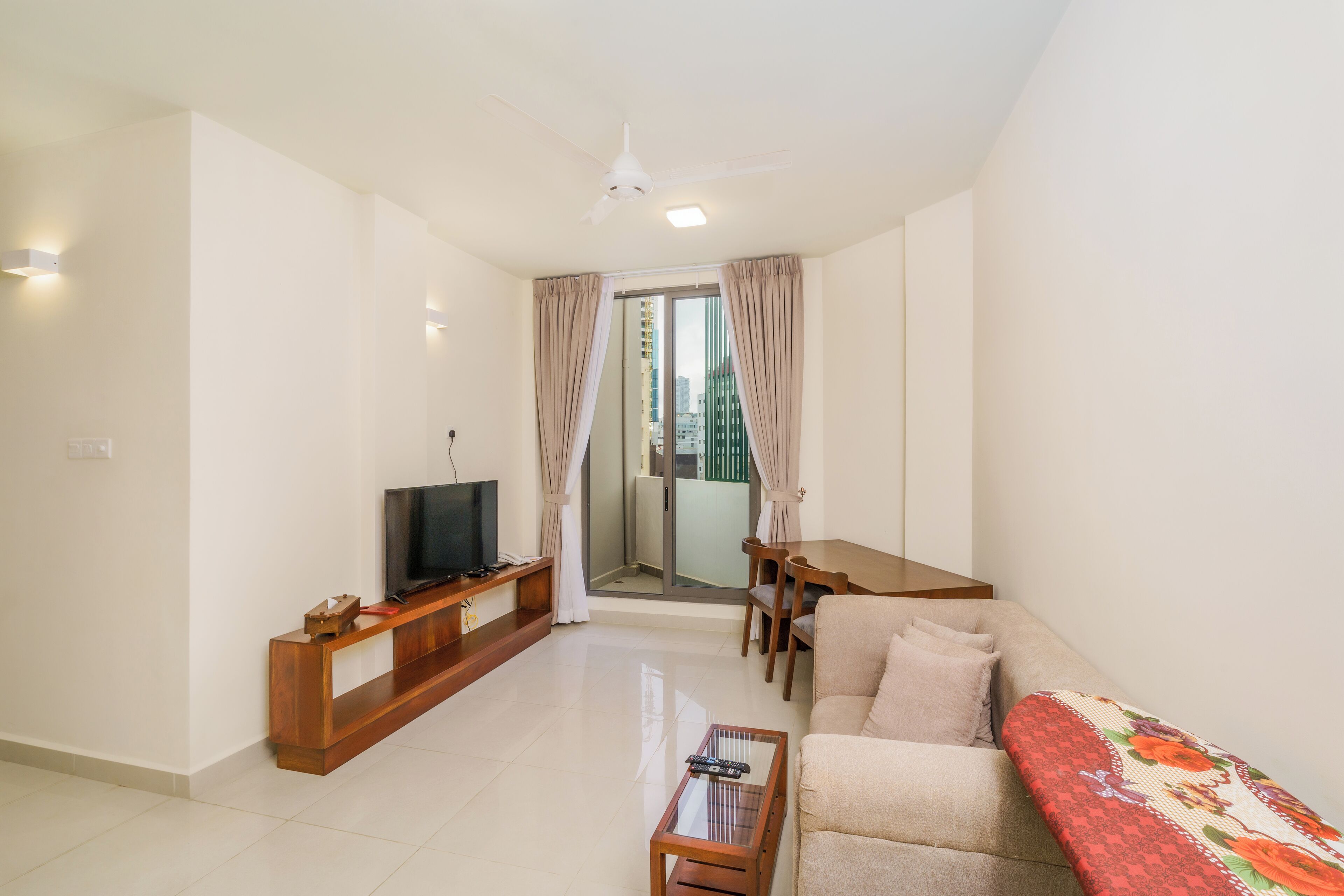 A modern living room with a flat-screen TV, wooden furniture, a glass coffee table, and a balcony with curtains.