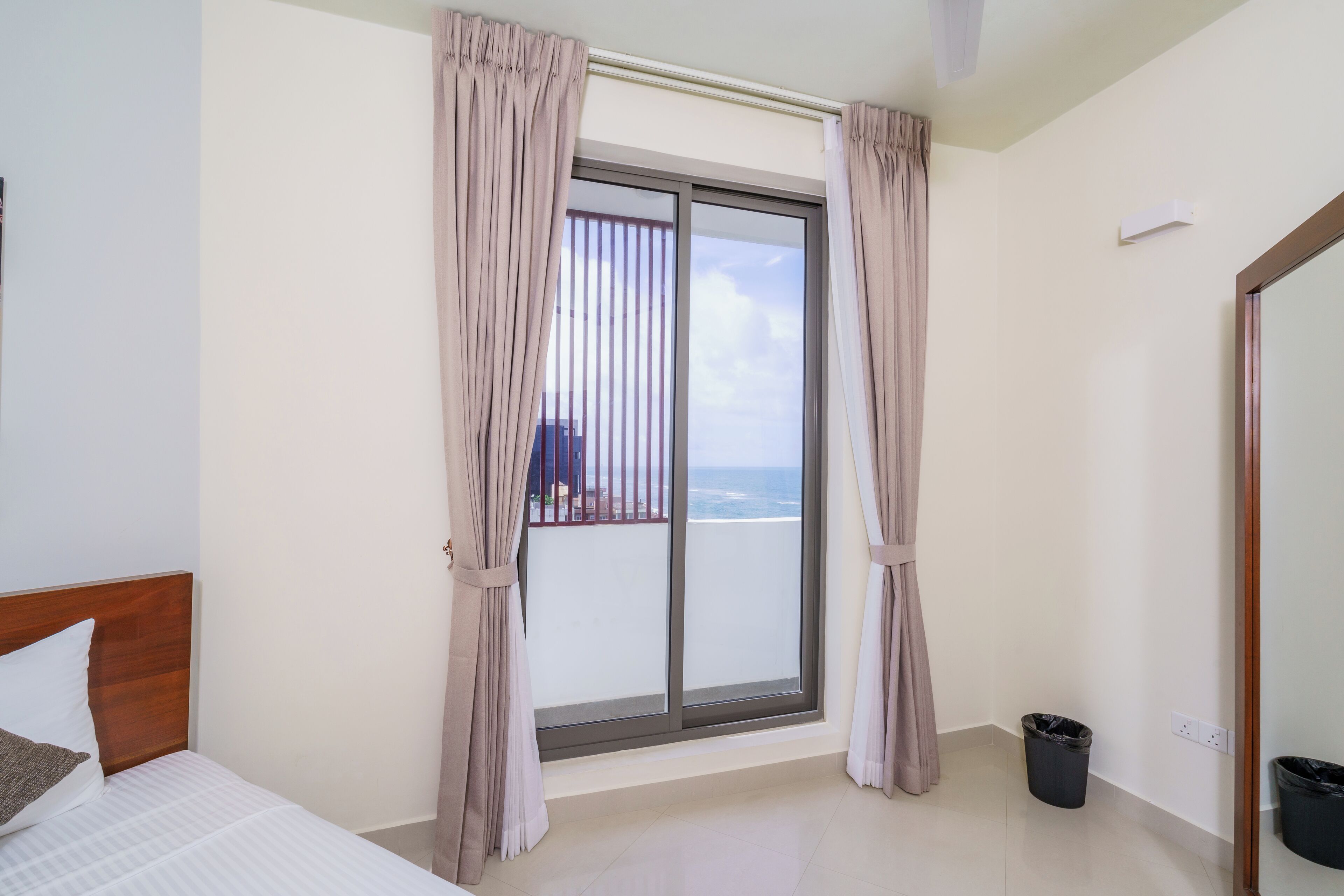 A hotel room with a large sliding glass door to the balcony, a bed with white bedding, and a small trash bin.