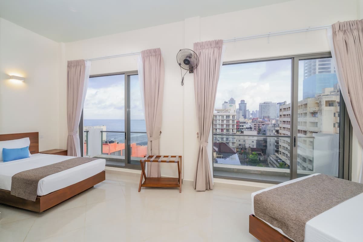 Quarto de hotel moderno com uma cama grande, uma cama menor, um ventilador e vista da cidade através de grandes janelas.