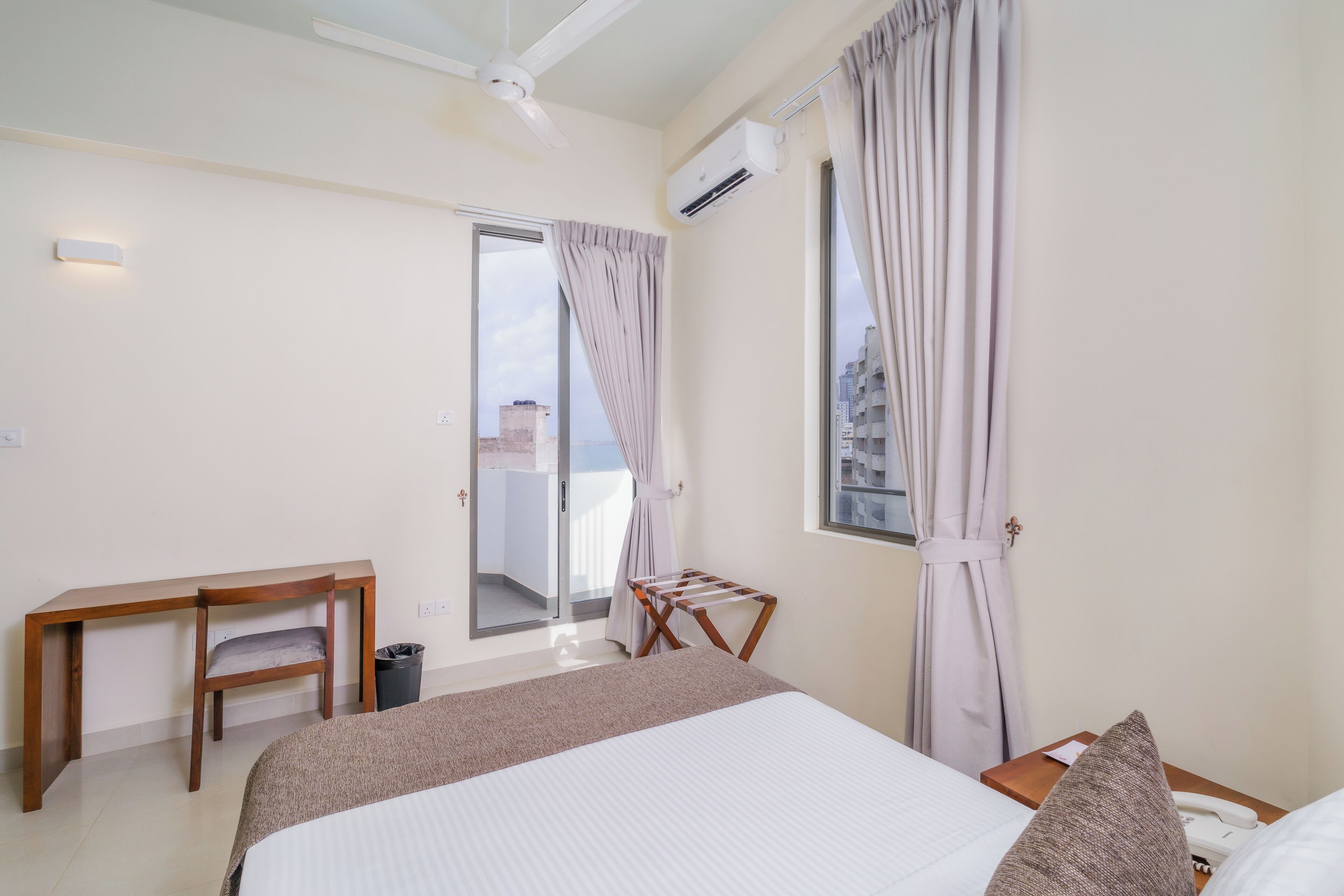 Une chambre d’hôtel avec un lit, un bureau, une chaise, un étendoir et une vue sur la ville par la fenêtre.