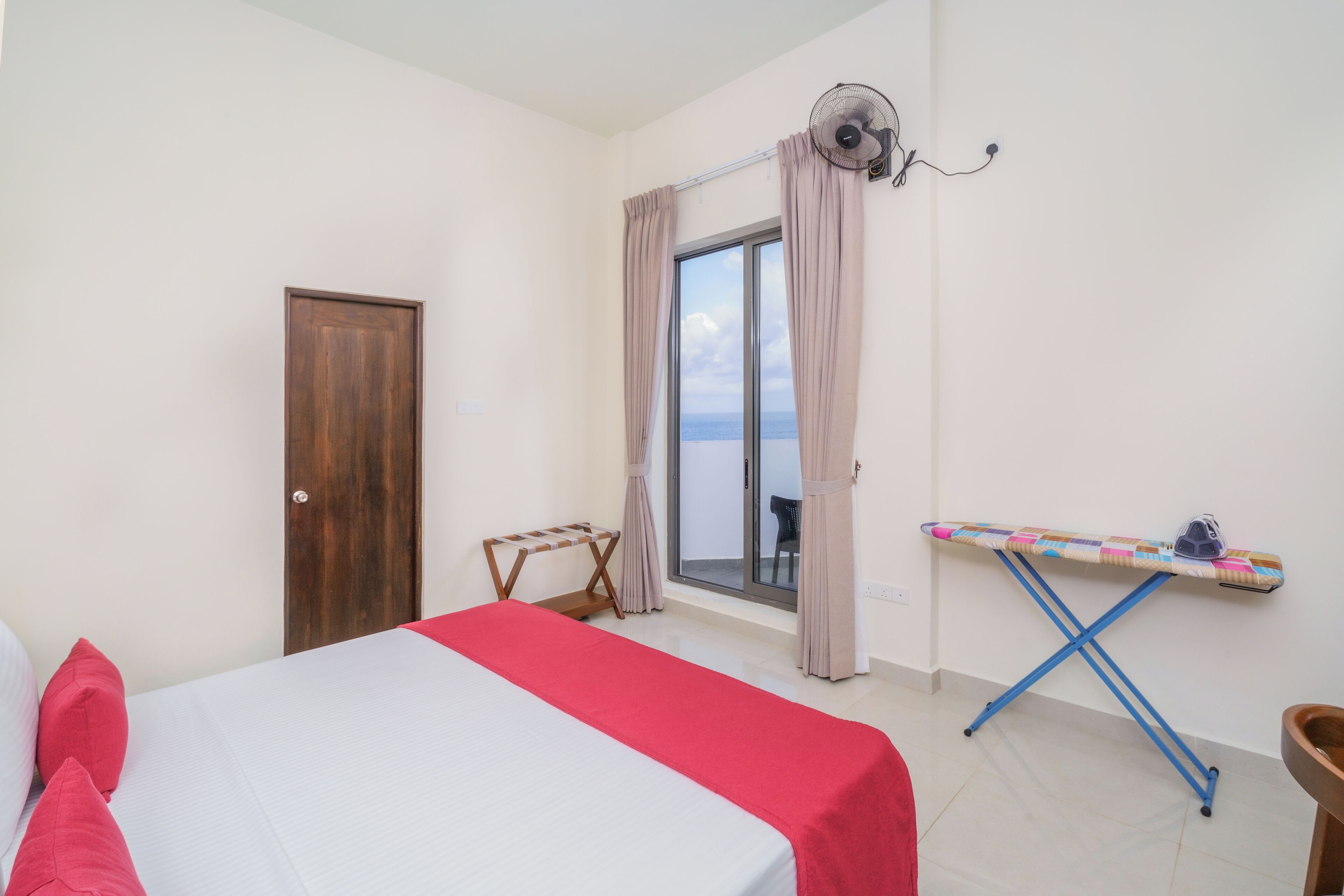 A hotel room with a bed, a window with curtains, a fan, and an ironing board.