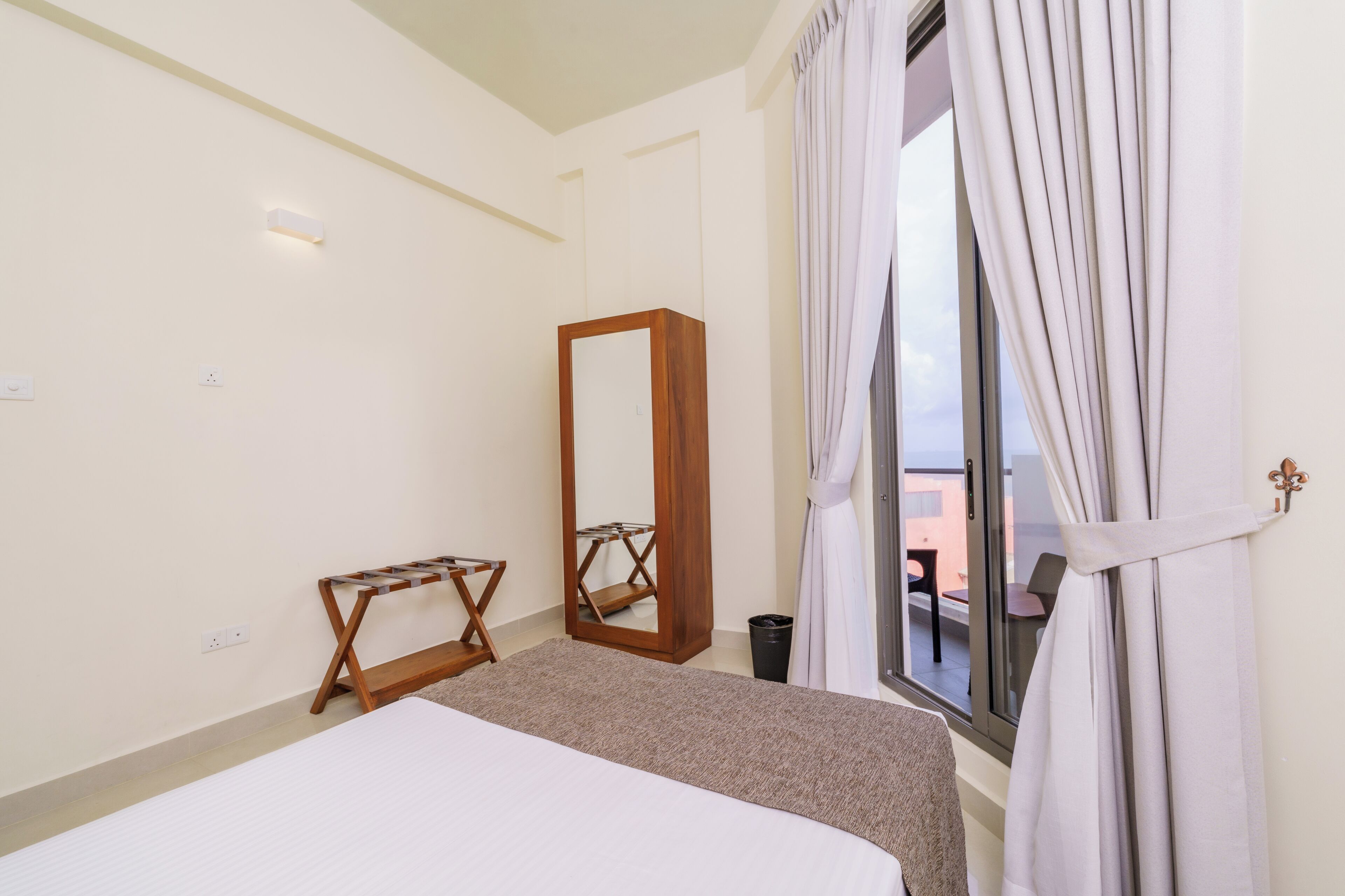 Une chambre d’hôtel avec un lit, un banc en bois, un miroir et une fenêtre avec des rideaux.