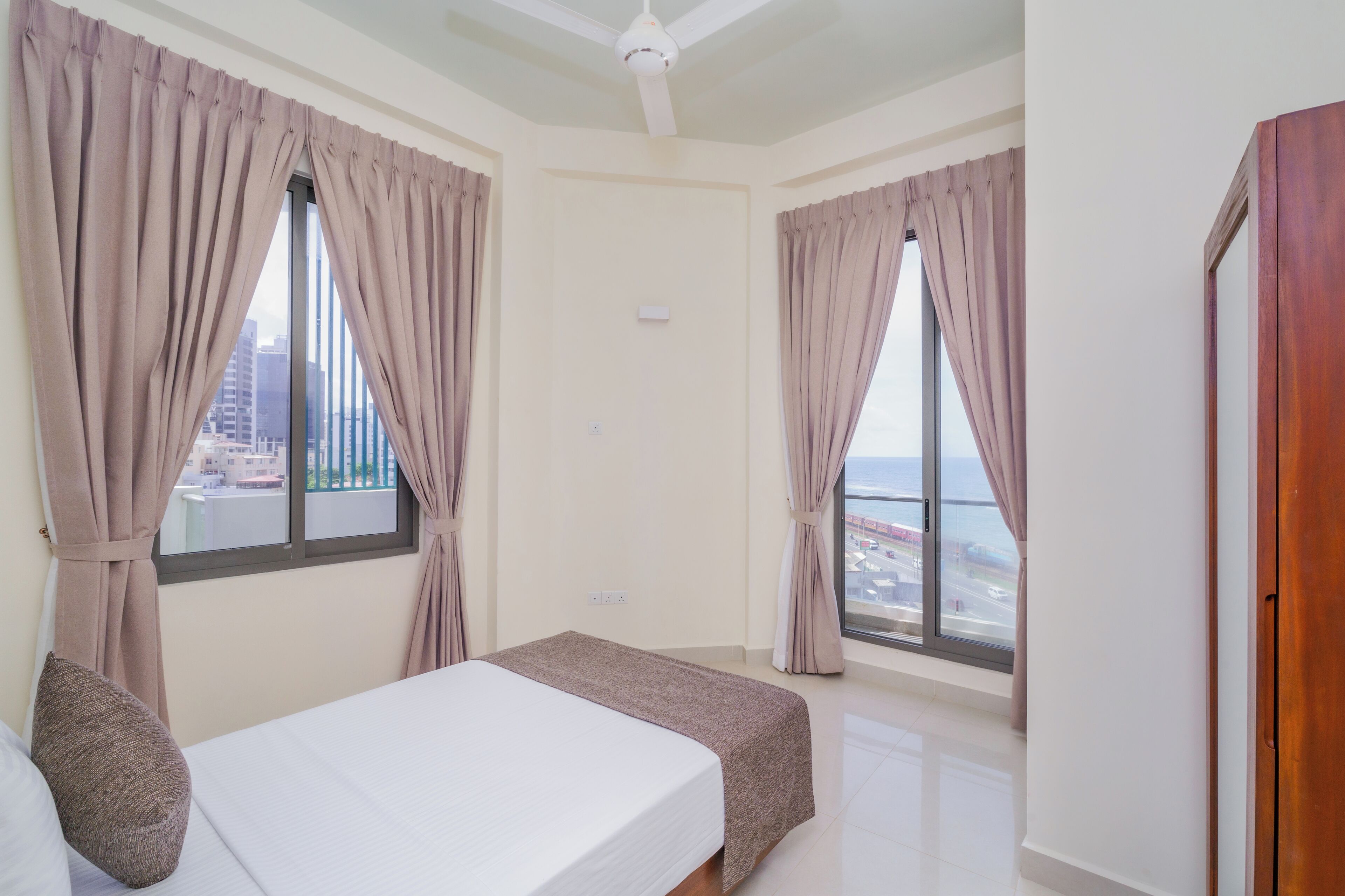 Una habitación de hotel con cama, dos ventanas con cortinas, un ventilador de techo y vistas a la ciudad.