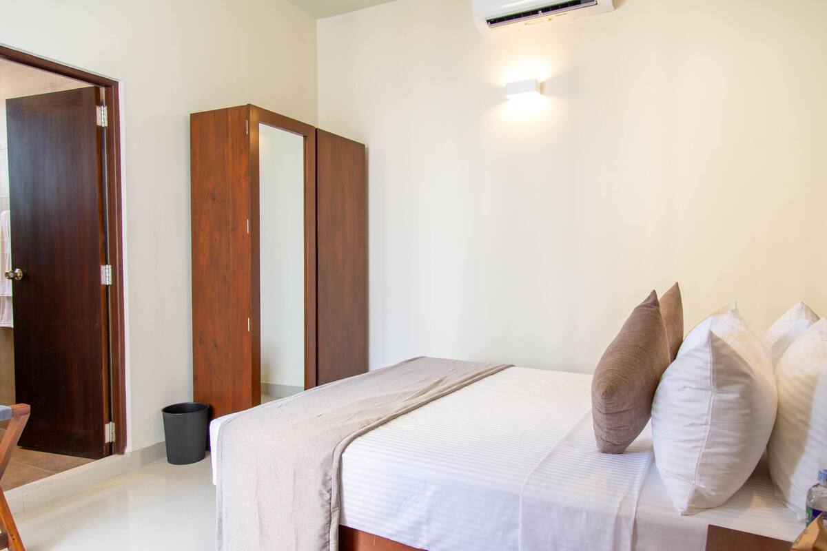 Quarto com cama, almofadas, guarda-roupa de madeira, lixeira e ar condicionado fixado na parede.