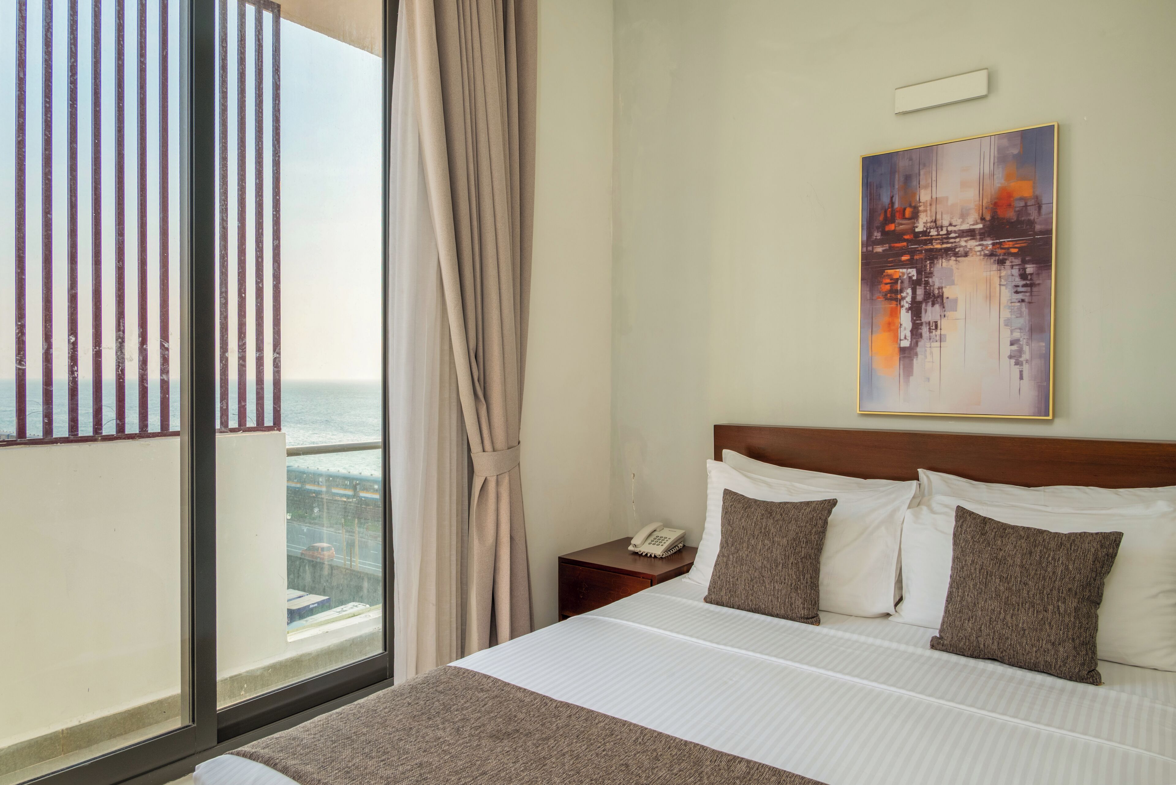 A hotel room with a large window, a bed with white bedding, a nightstand with a phone, and a painting on the wall.
