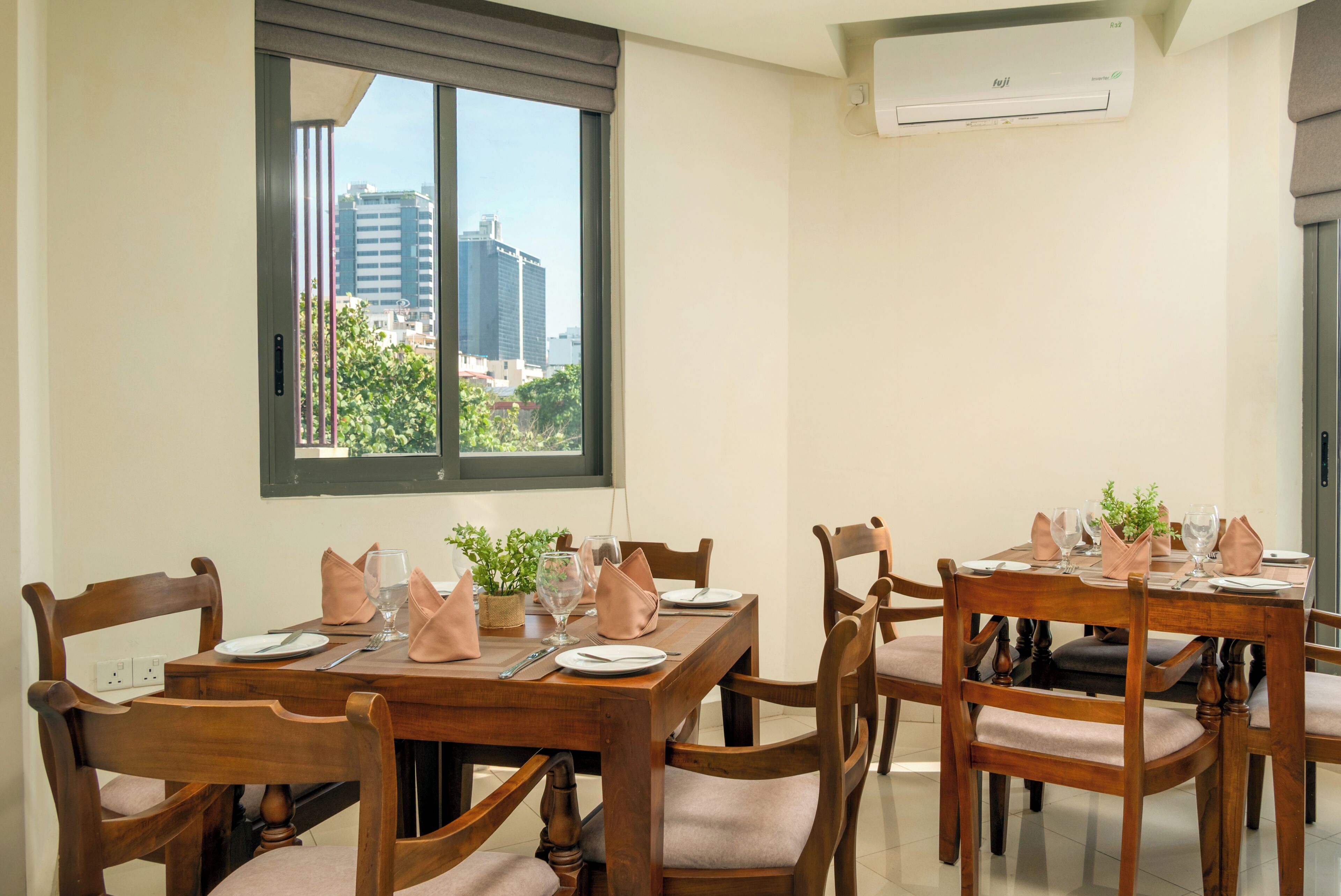 Zona pranzo con tavolo apparecchiato per quattro, sedie in legno, una finestra con vista sugli edifici e un condizionatore d'aria a parete.