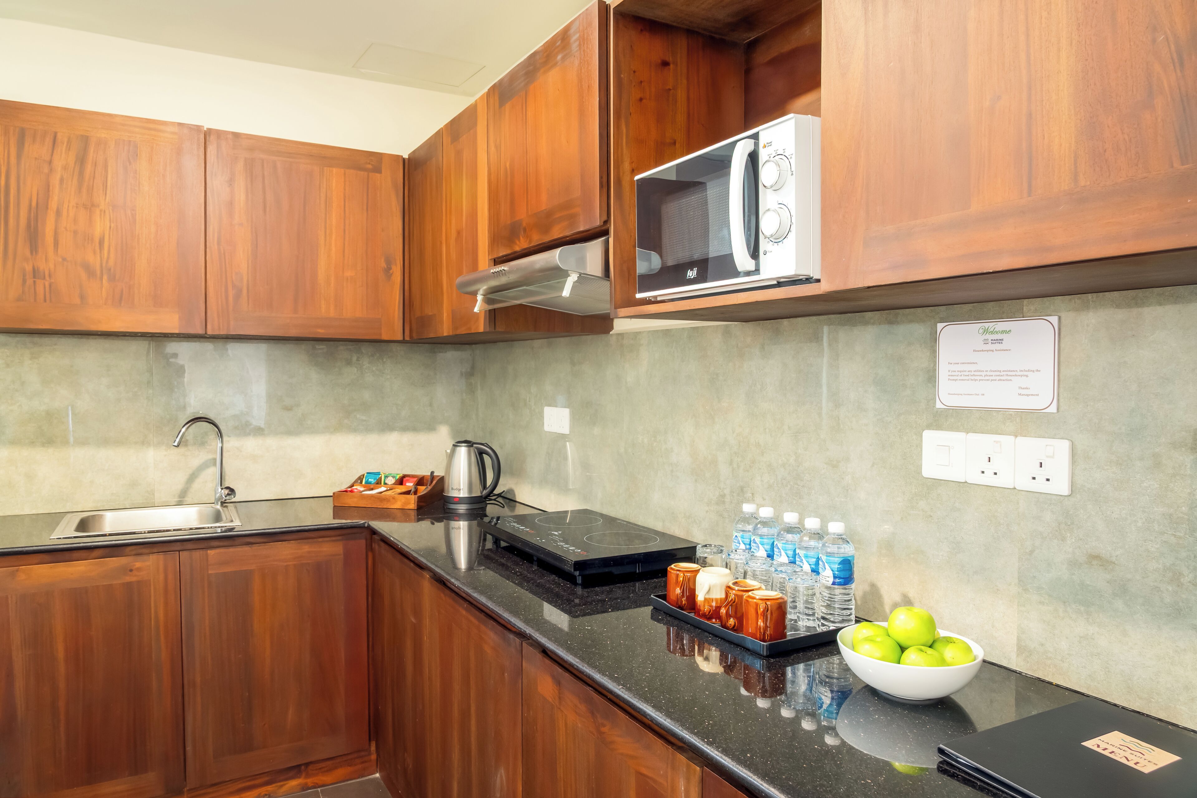 Una cocina con armarios de madera, microondas, hervidor de agua, cafetera, encimera negra, un bol de manzanas y botellas de agua.