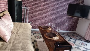 A room with a brick wall, a bed with beige bedding, a wooden coffee table with plates and glasses, and a TV mounted on the wall. - Studio Neuf à Maillane (Maillane)