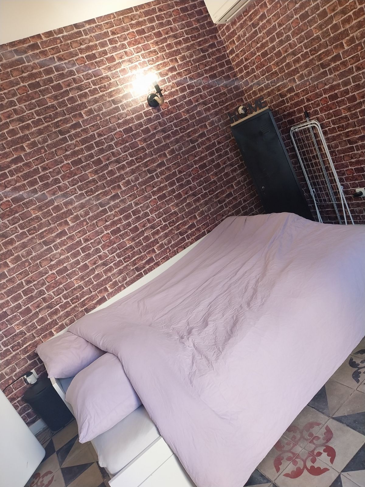 A room with a brick wall, a bed with a light purple bedspread, and a small table with a white chair.