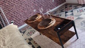 A room with a brick wall, a flat-screen TV mounted on the wall, a wooden table with woven placemats and wine glasses, a beige blanket, and patterned floor tiles. - Studio Neuf à Maillane (Maillane)