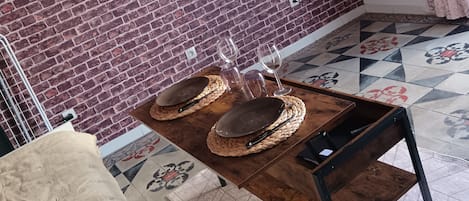 A room with a brick wall, a flat-screen TV mounted on the wall, a wooden table with woven placemats and wine glasses, a beige blanket, and patterned floor tiles.
