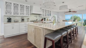 Une cuisine spacieuse avec des meubles blancs, un grand îlot central doté de tabourets, et une vue sur l’océan grâce à de grandes fenêtres.
