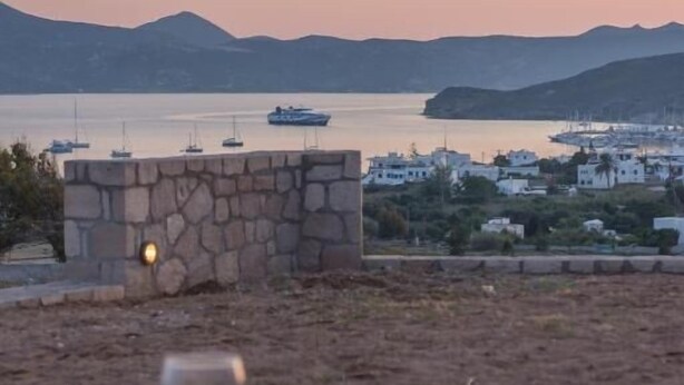 Vista costeira com muro de pedra, uma mesa com um copo de vinho tinto e barcos ao fundo.