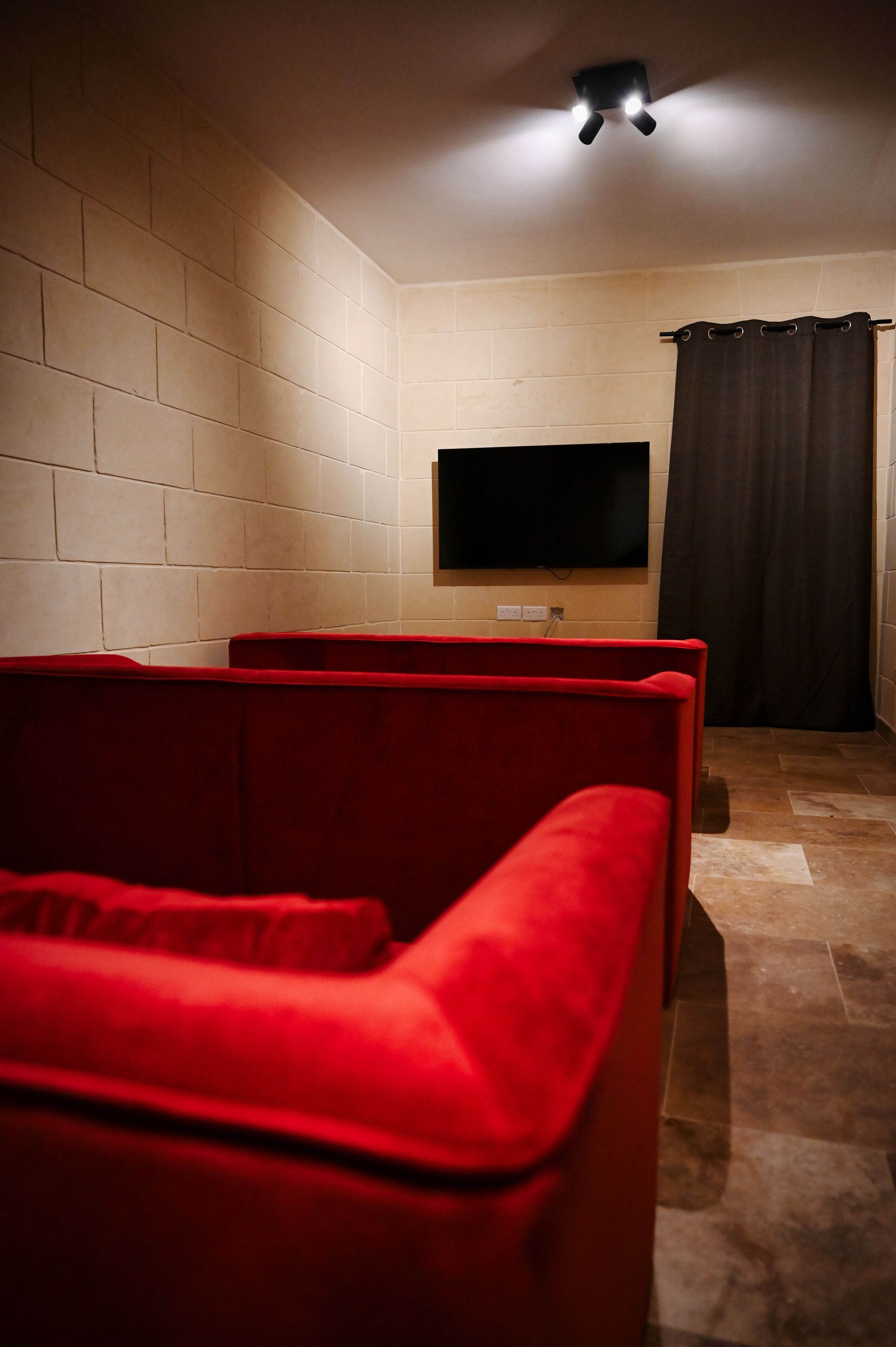 A room with a flat-screen TV mounted on a brick wall, a red couch, and a dark curtain.