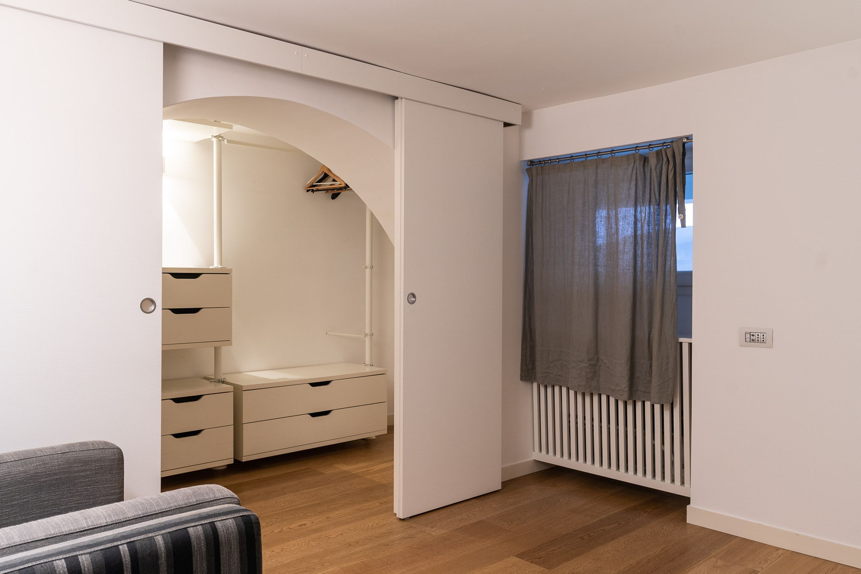 A room with a white wardrobe, a grey sofa, and a radiator.