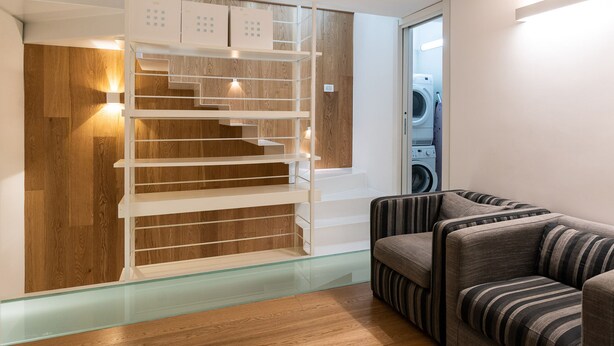 A modern interior with a wooden floor, a striped sofa, a glass staircase, and a washing machine in the background.