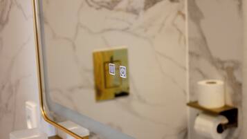 Un baño con paredes de mármol, un espejo, un dispensador de papel higiénico y un botón para la descarga.