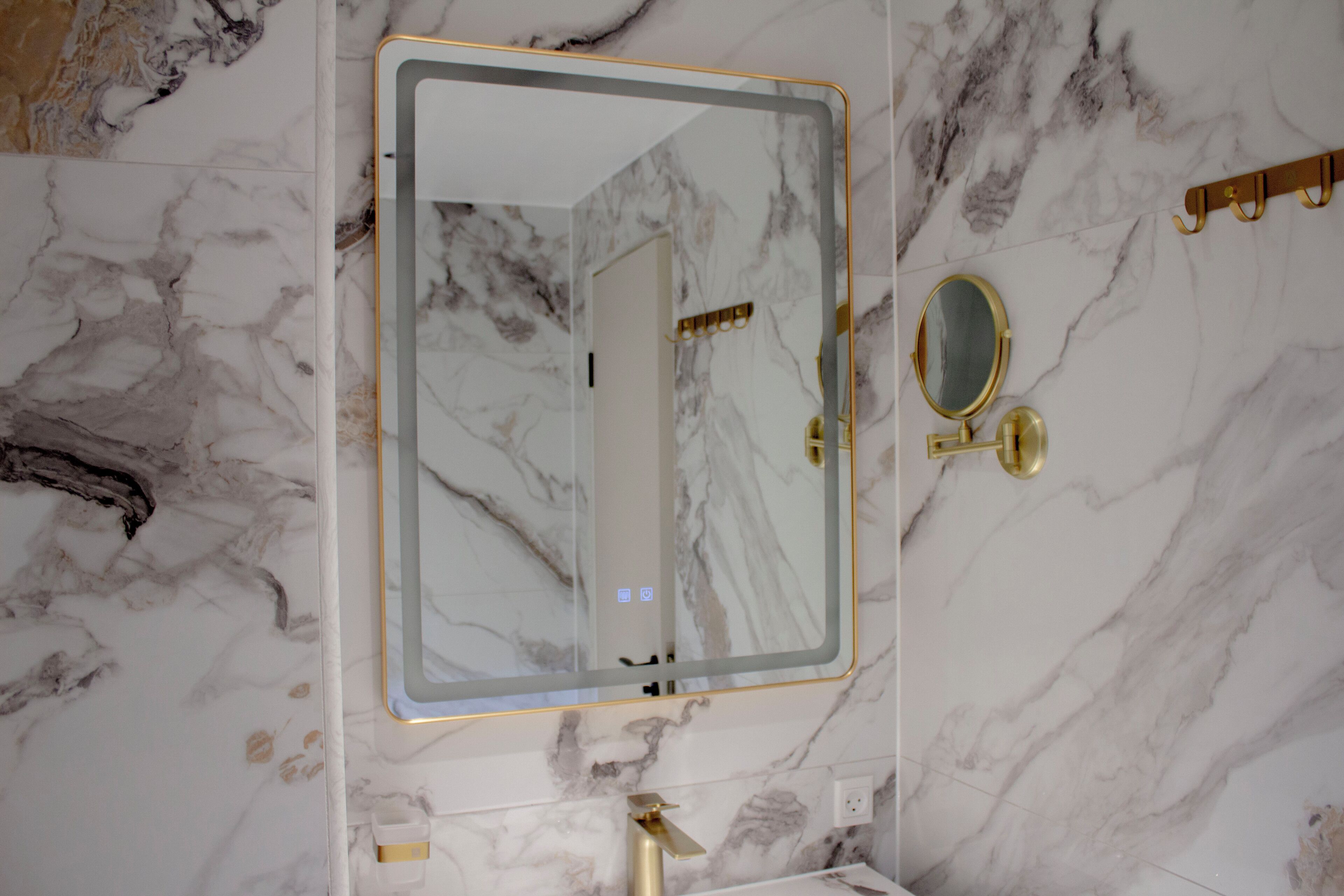 Ein Badezimmer mit Marmorwand, einem großen Spiegel mit goldener Rahmung, einem Wasserhahn in Gold und einem Handtuchhalter aus Gold.