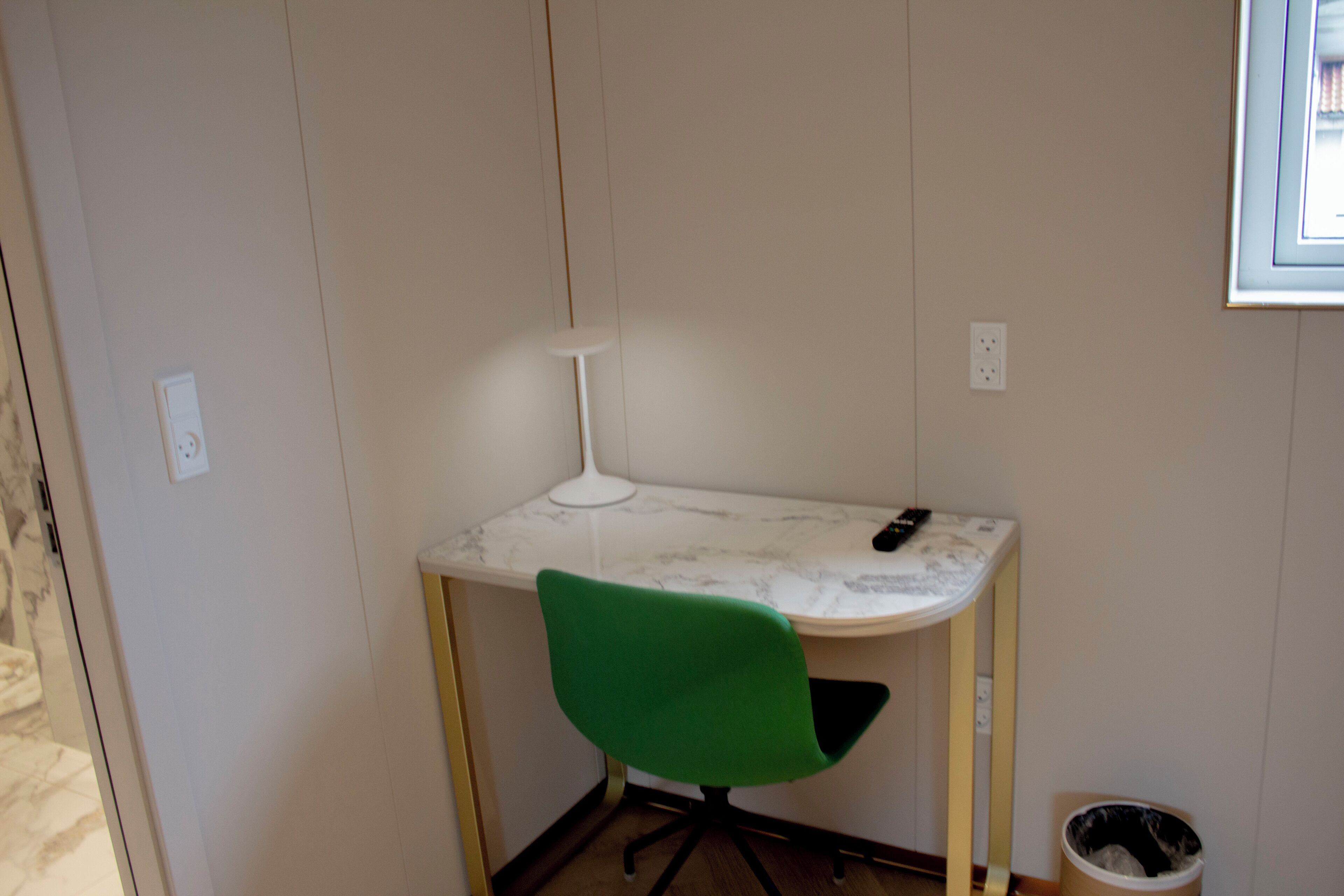 Un espacio de oficina pequeño y privado con un escritorio blanco, una silla verde, una lámpara blanca y un cubo de basura.
