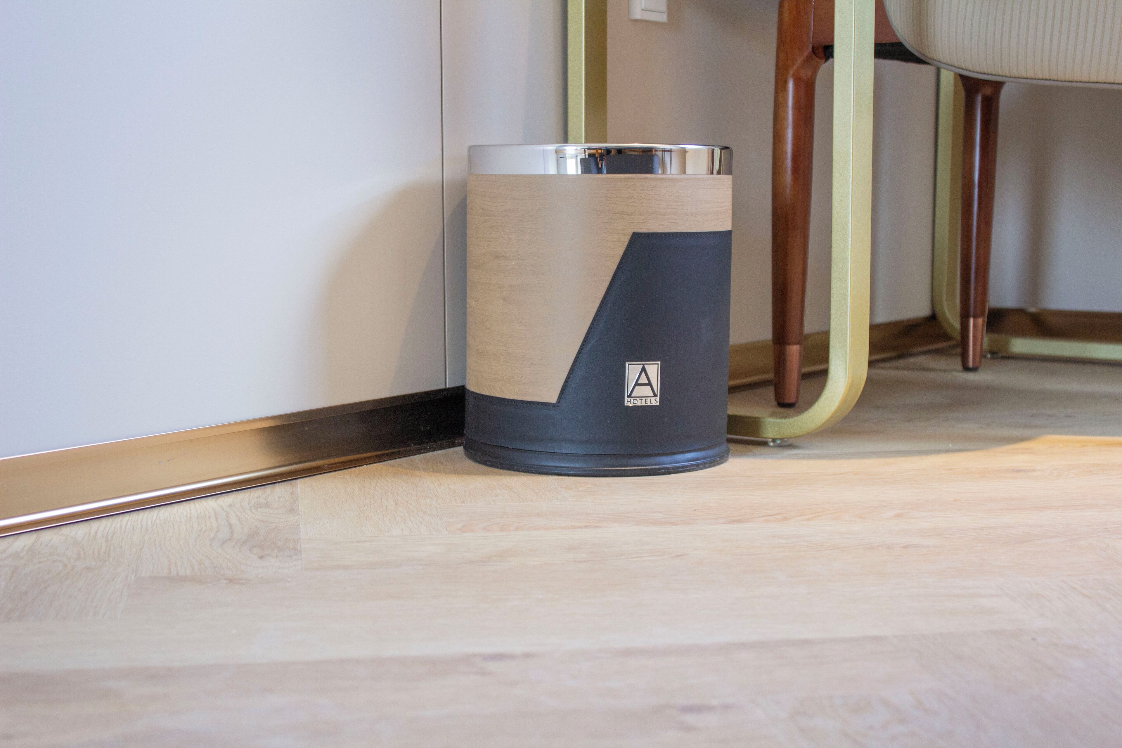 A cylindrical trash bin with a wooden and metallic design, placed on a wooden floor near a wall and a chair.