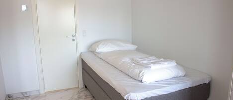 Una habitación pequeña con cama individual, un lecho gris, ropa blanca y un suelo con estampado de mármol, y una puerta cerrada.
