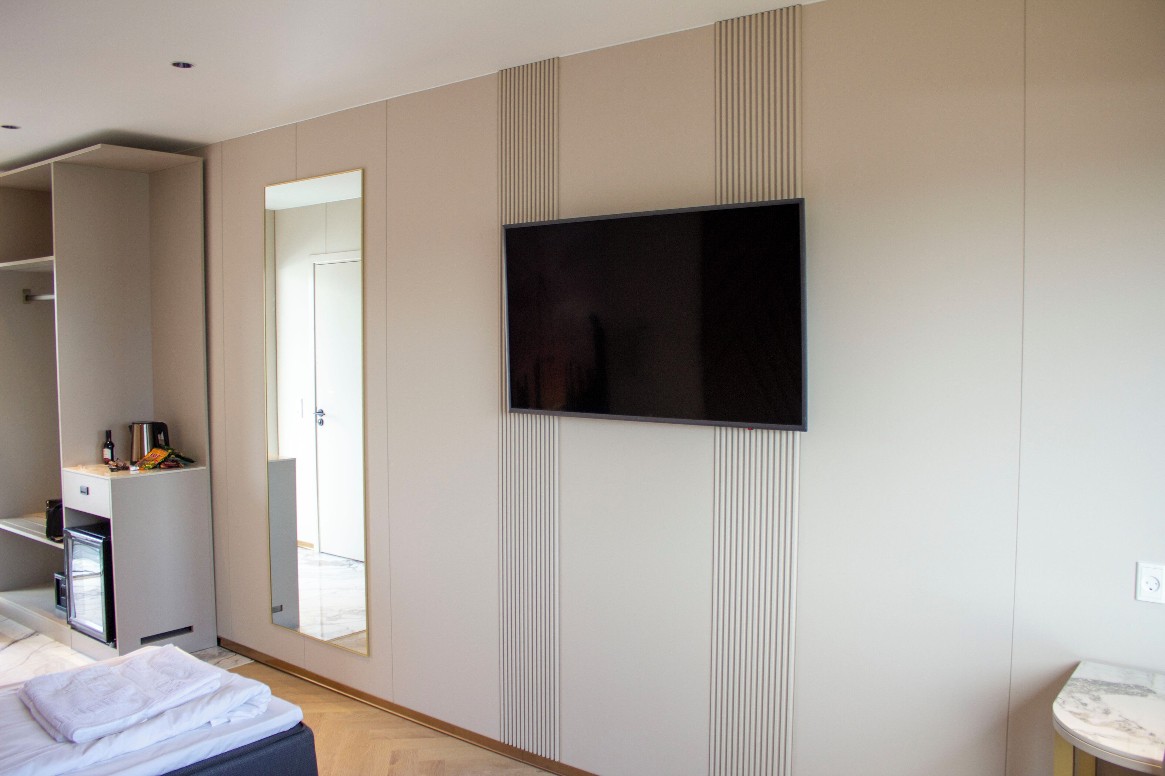 A modern hotel room with a large bed, a flat-screen TV mounted on the wall, a built-in wardrobe, and a small bathroom area.