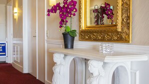 Un pasillo con una consola de mármol blanco, una orquídea en maceta, un espejo con marco dorado y un cubo de cristal transparente.