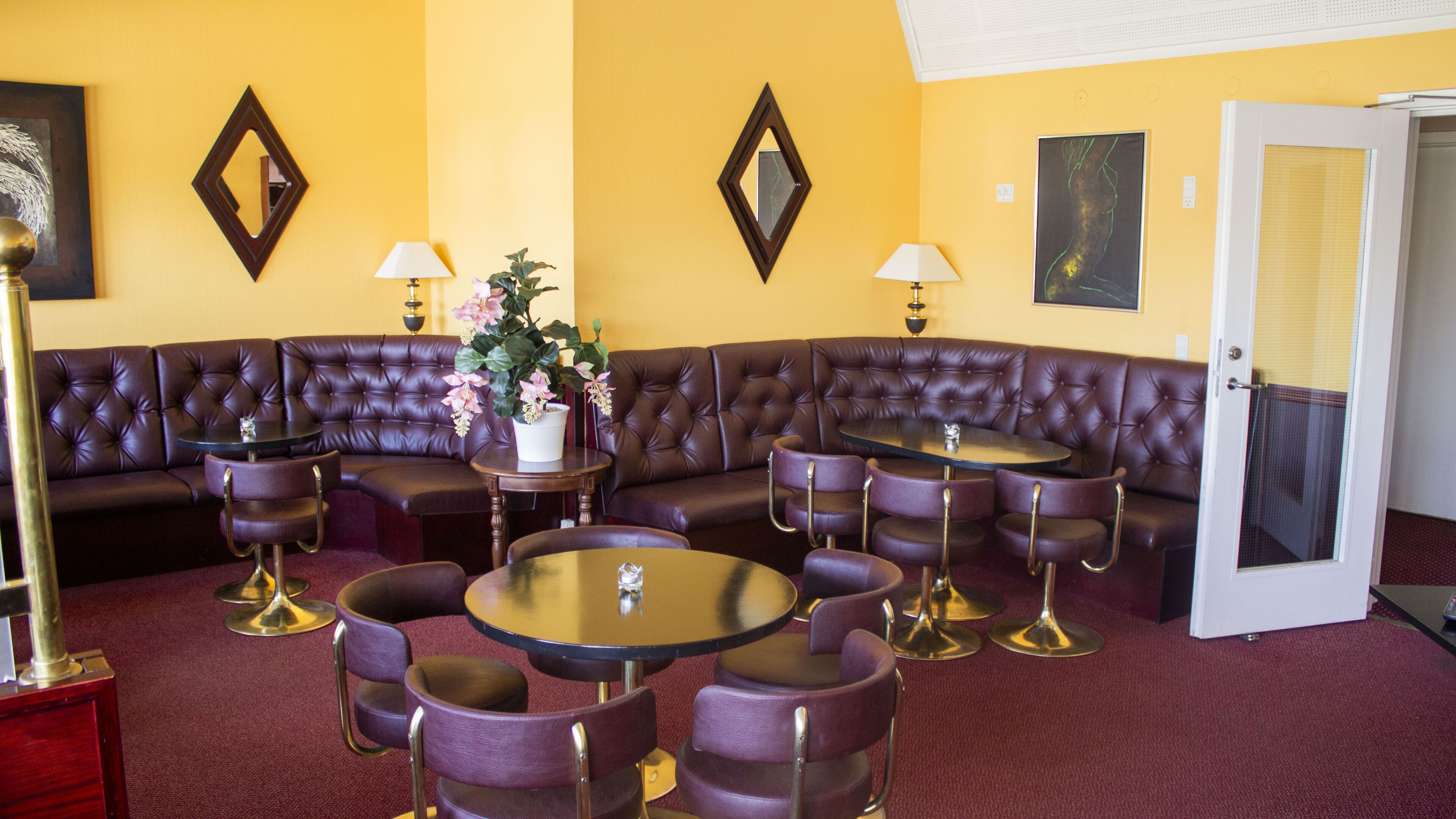 A room with a large L-shaped leather sofa, multiple round tables, and purple chairs. There are two wall-mounted lamps and a potted plant.