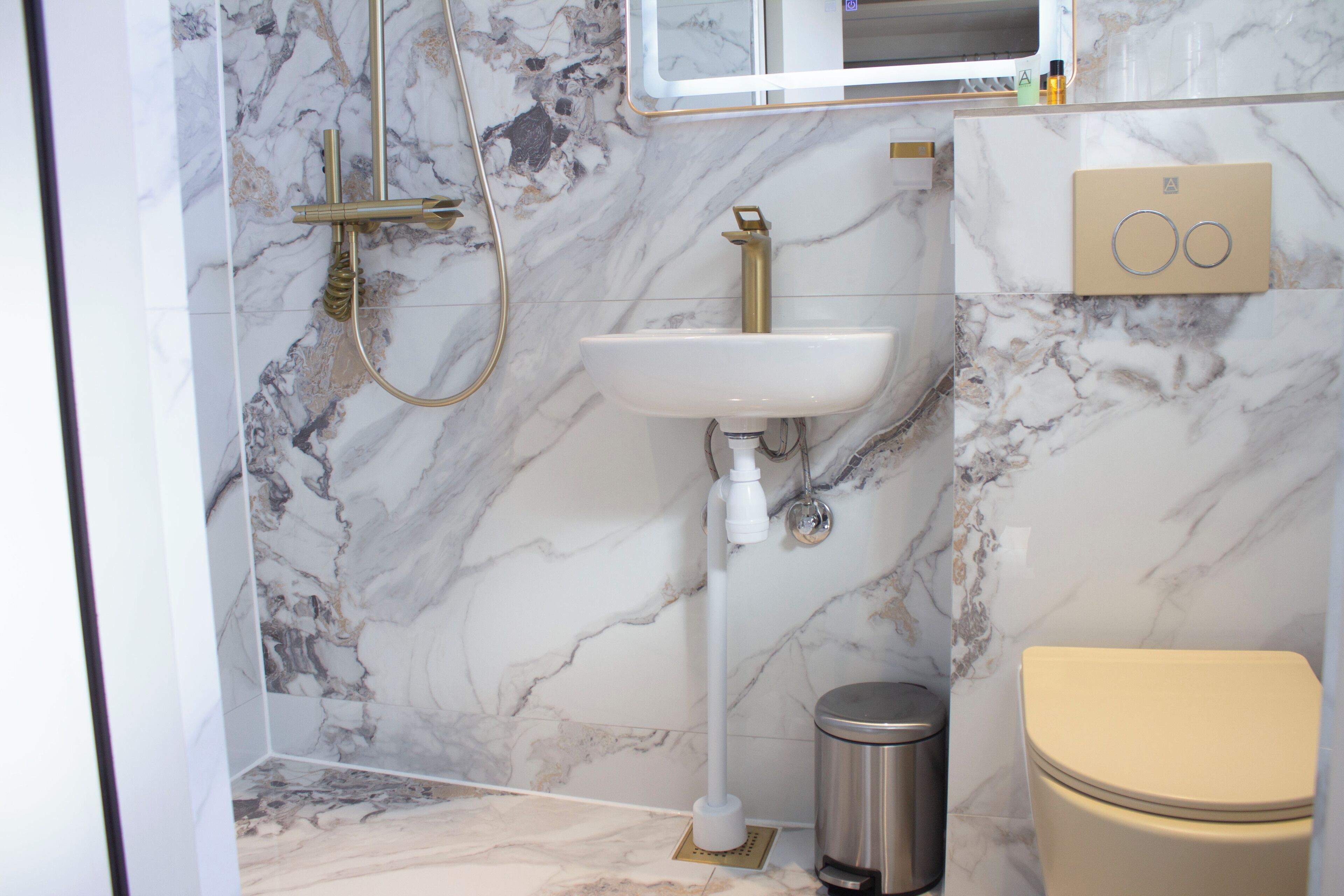 A bathroom with a marble wall, a white sink, a gold faucet, a toilet, and a trash can.