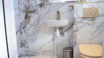 A bathroom with a marble wall, a white sink, a gold faucet, a toilet, and a trash can.