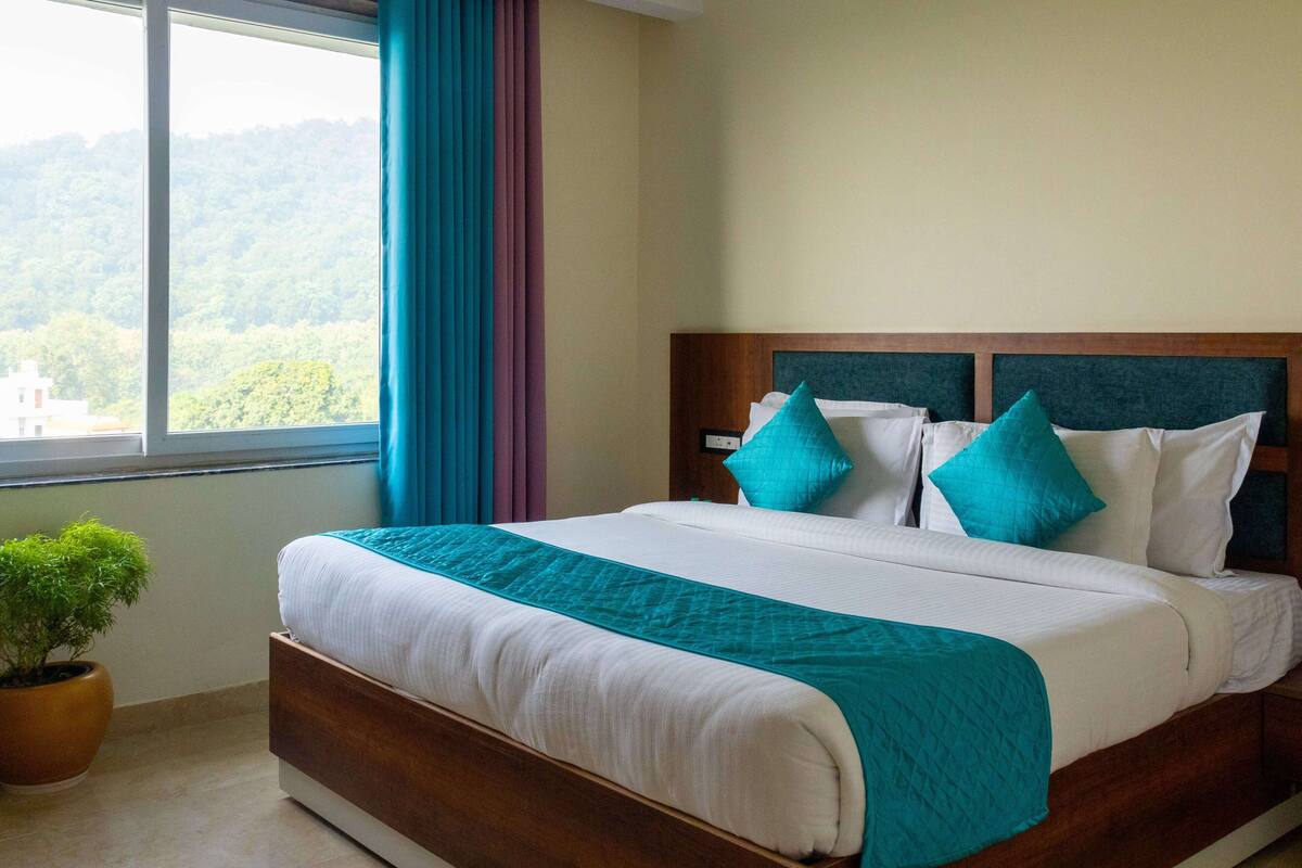 A hotel room with a large bed, a view of the outdoors through a window, and a potted plant on the floor.