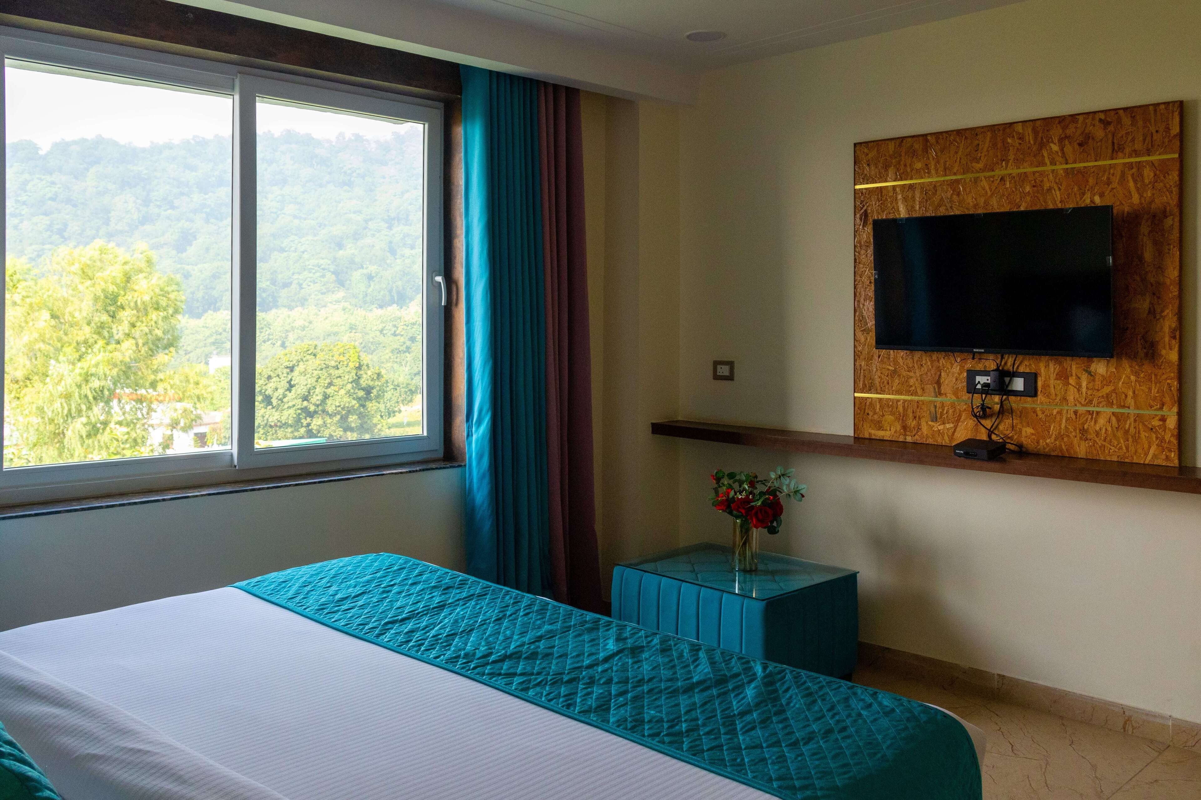 Una habitación de hotel con una cama grande, un televisor instalado en un panel de madera, una mesita con un jarrón de flores y una ventana con cortinas.