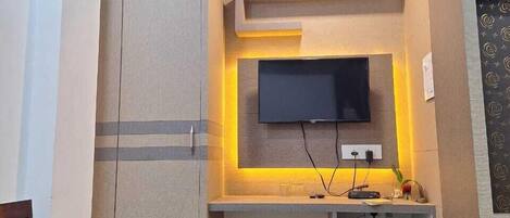 A compact living space with a flat-screen TV mounted on the wall, a small cabinet below, and a bed with a headboard.