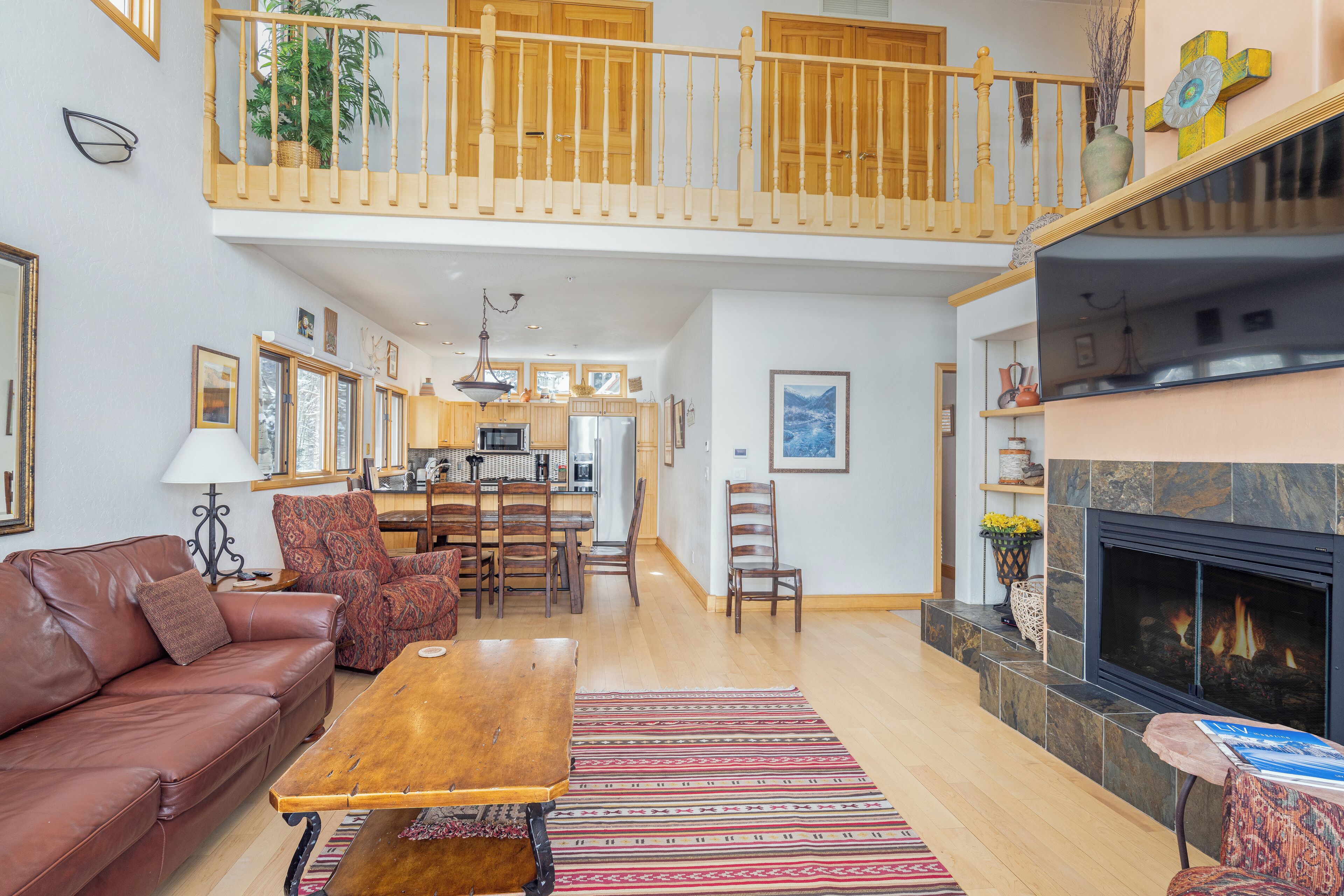 A spacious living area with a high ceiling, a fireplace, a dining area, and a kitchen in the background.