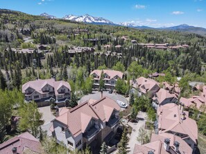 A residential area with multiple houses, some with red roofs, surrounded by dense forest and mountains in the background.