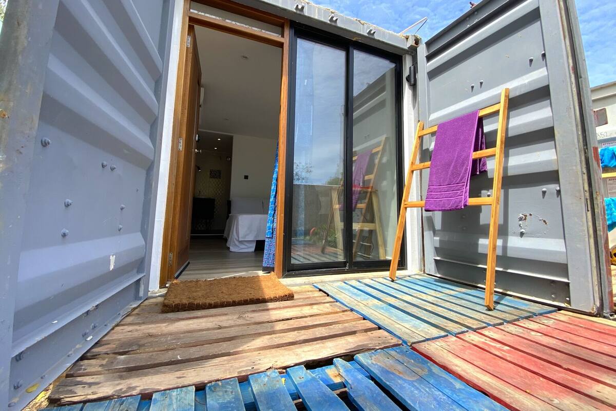 Un interior moderno y funcional, con una puerta corredera de vidrio que da a una habitación, una pequeña terraza de madera y una toalla morada sobre una escalera de madera.