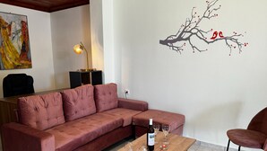 A living room with a pink sectional sofa, a wooden coffee table with wine glasses and a bottle, and a wall decal of a tree with birds. - Skiathos Modern Garden Escape Apt (Skiathos)