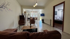 A living room with a brown sofa, a dining area with chairs, a TV mounted on the wall, and a kitchen in the background. - Skiathos Modern Garden Escape Apt (Skiathos)