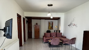 A living room with a sectional sofa, a dining table with chairs, a flat-screen TV mounted on the wall, and a decorative wall piece. - Skiathos Modern Garden Escape Apt (Skiathos)